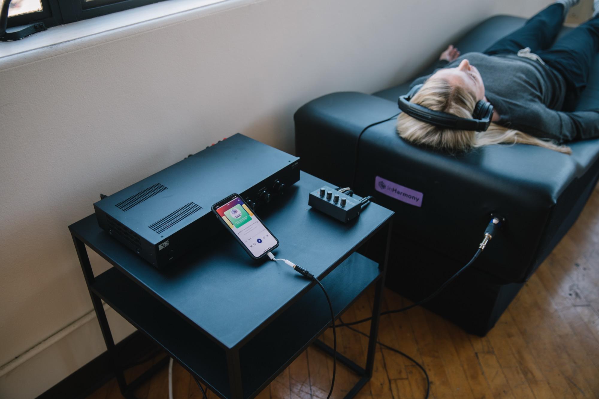 A person lying on the vibroacoustic table with headphones on, the amplifier and phone on a side table beside them