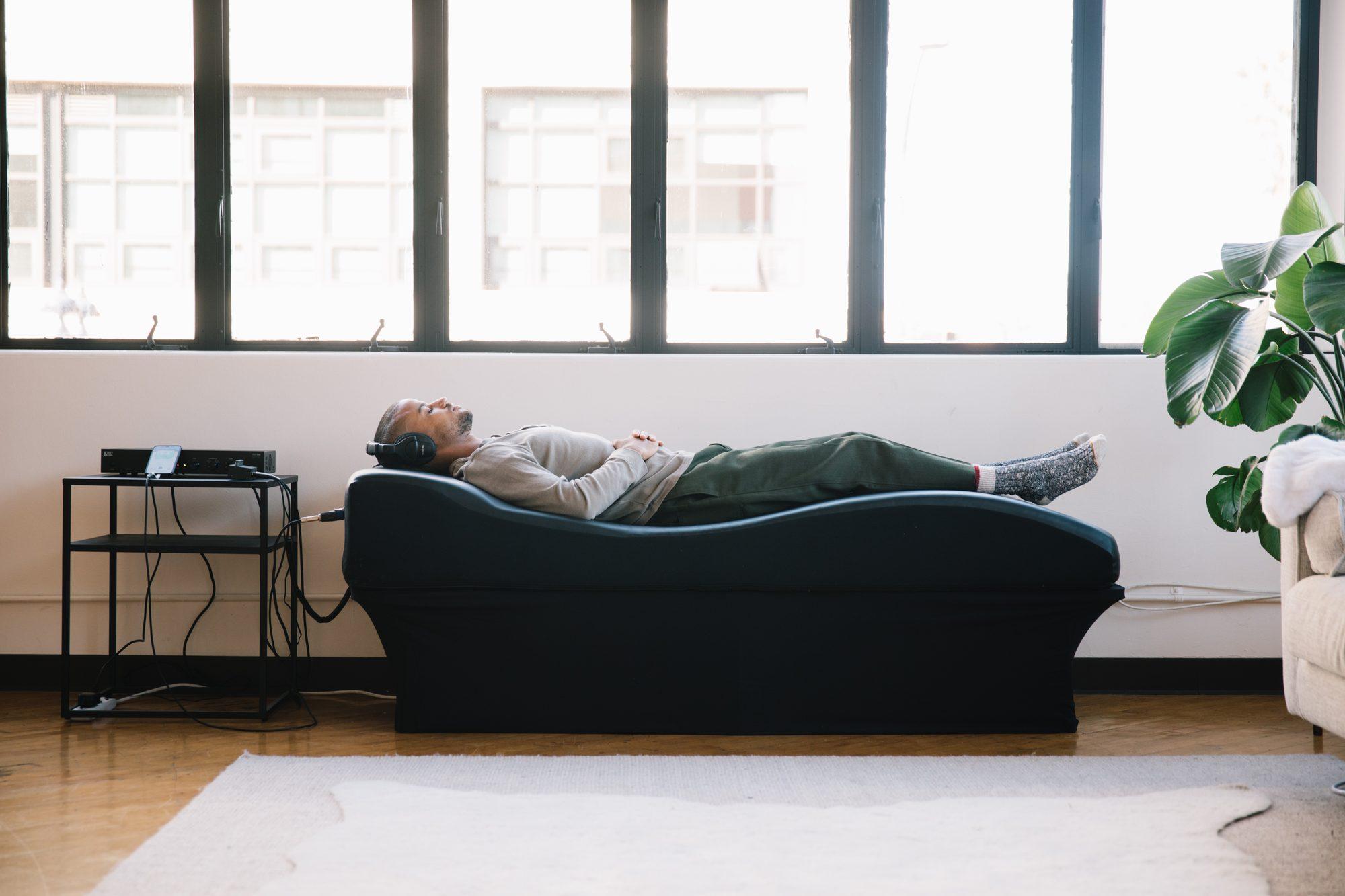 A person lying fully clothed on the vibroacoustic table with headphones on, in a bright room by a window