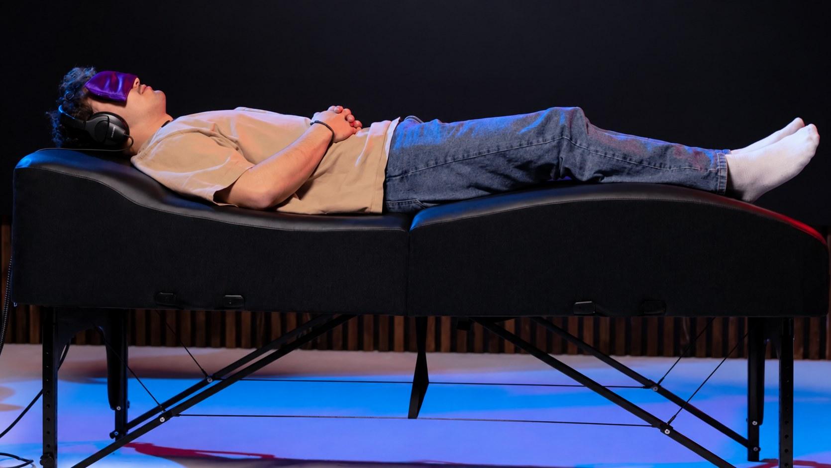 A guest under a blanket with a sleep mask and headphones on the table at a pop-up event, lit in violet and gold